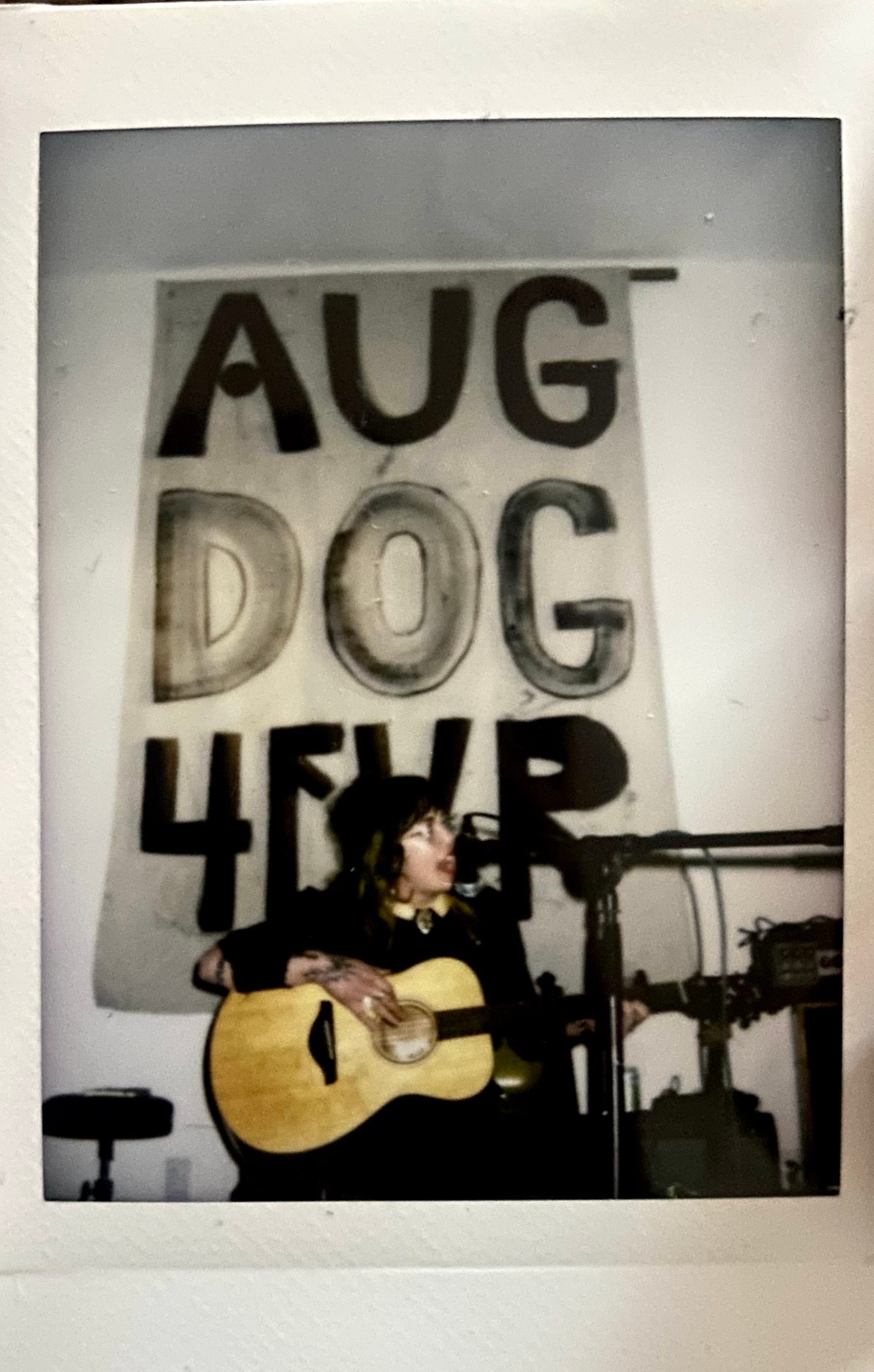 Caitlin Angelica playing acoustic guitar wearing black in front of a banner that reads “AUG DOG 4EVR,” photo courtesy the artist